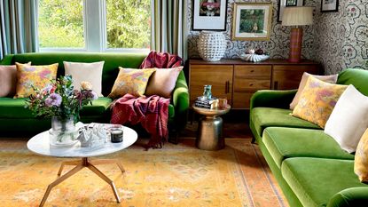 An eclectic-style living room with two green, velvet couches in a caddy-corner to each other with a yellow-orange rug in between. There is a window, blue and white wallpaper, and a white marble coffee table in the room.