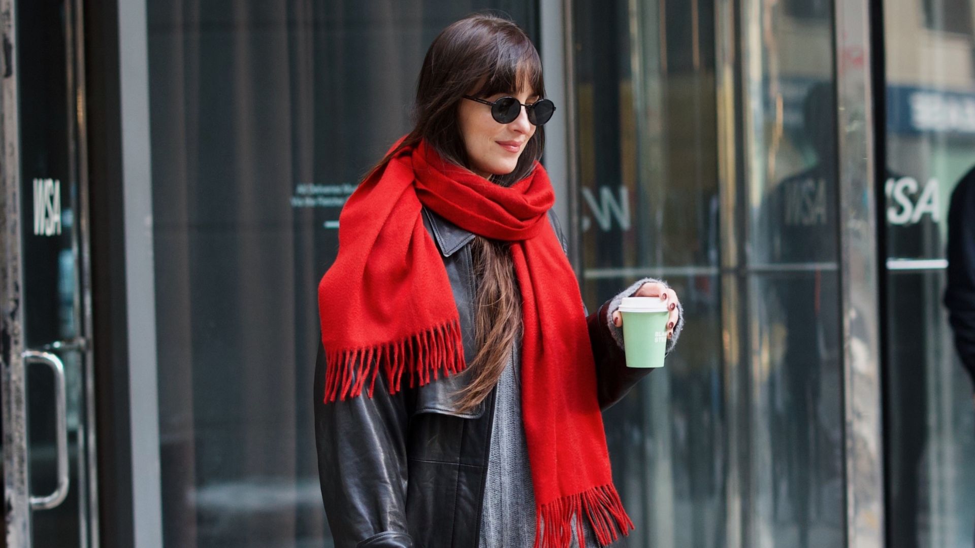 Dakota Johnson walks outside wearing black straight-leg jeans with loafers, a grey knit, a black leather jacket, a red scarf and a leopard print bag.