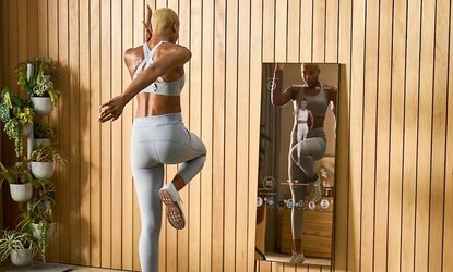 woman working out using the Lululemon Mirror, which is leant up against a wood panelled wall, with plants in the corner