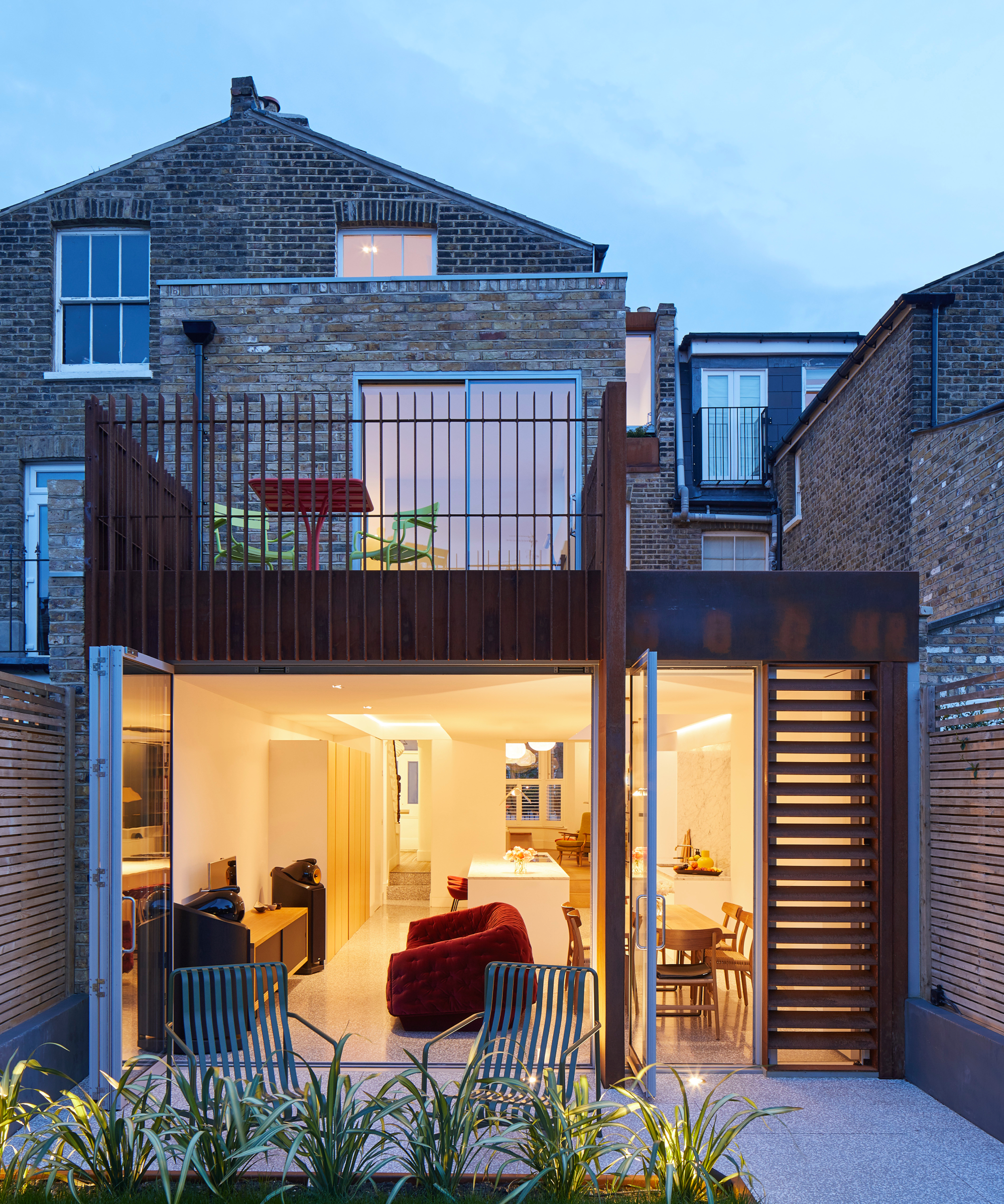 corten clad wraparound extension at night with bifold patio doors open showing open plan living room inside, outside there is a first floor terrace on top of the extension accessed via sliding doors in first floor room
