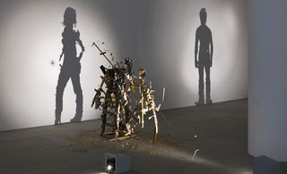 A closer view of one shadow sculpture made out of junk, metal, and wood. The light shines on the sculpture and casts a shadow on the wall. The shadow portrays a woman.