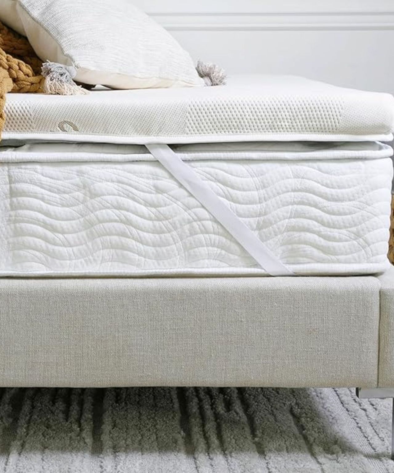 The Saatva Graphite Mattress Topper on a white mattress on a neutral bedframe, with a white cushion and an ochre knitted blanket on top. The wall is white, and the rug beneath is grey and striped.