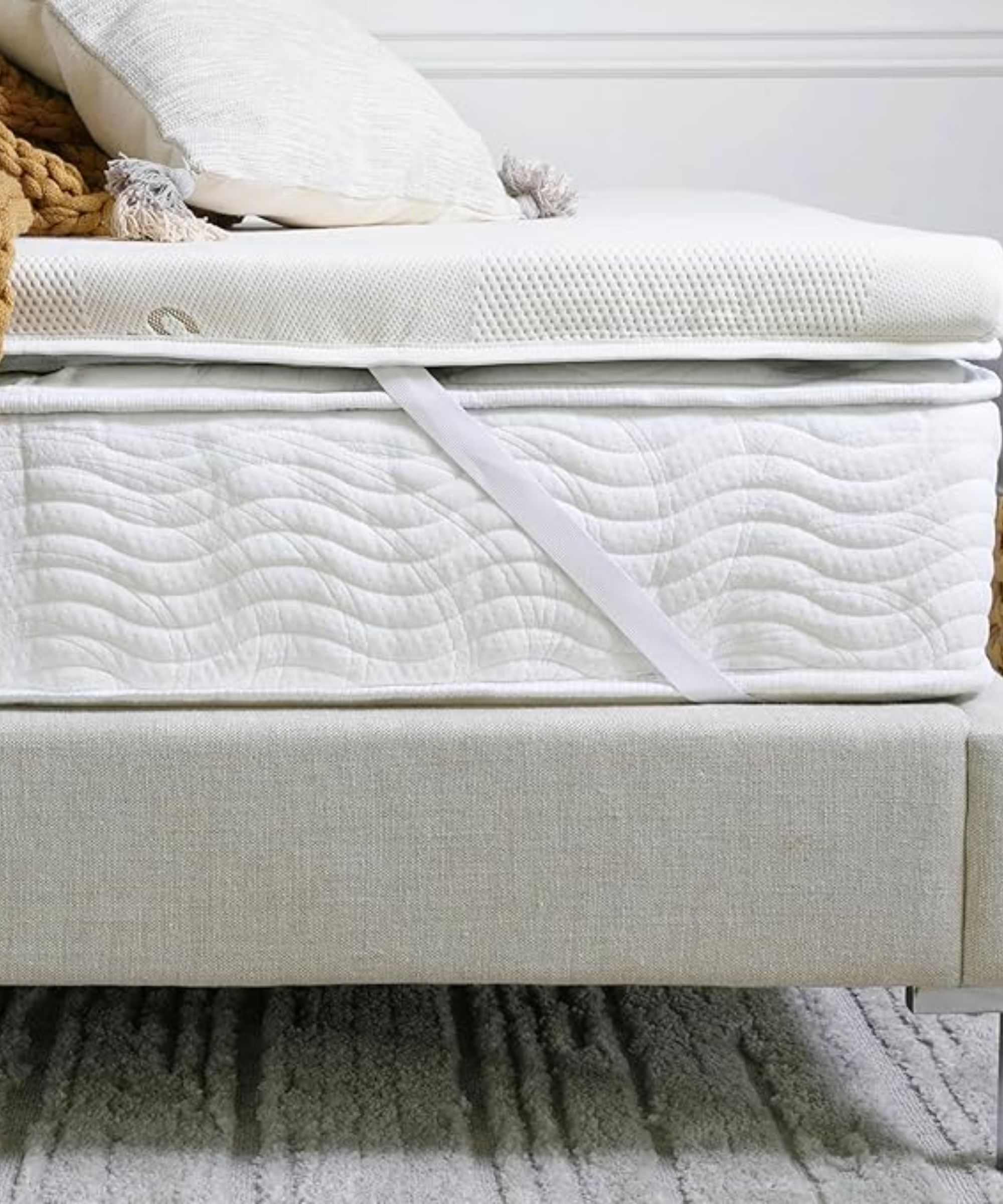 The Saatva Graphite Mattress Topper seen on a white mattress on a neutral bedframe, with a white cushion and the corner of an ochre colored knitted blanket on top. The wall behind is white, and the rug beneath is grey and striped.