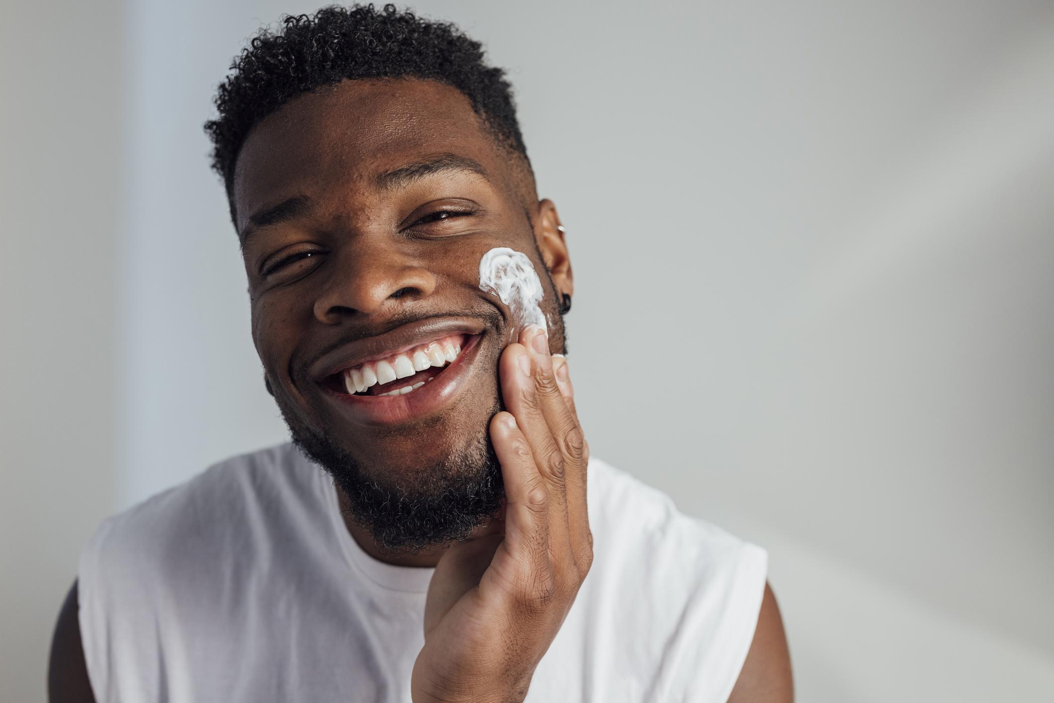  A close-up of a young man putting moisturiser on his cheek with his fingers while looking at the camera and smiling, he is looking after his skin. 