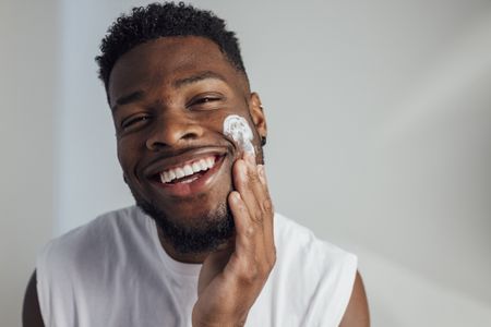 A close-up of a young man putting moisturiser on his cheek with his fingers while looking at the camera and smiling, he is looking after his skin.