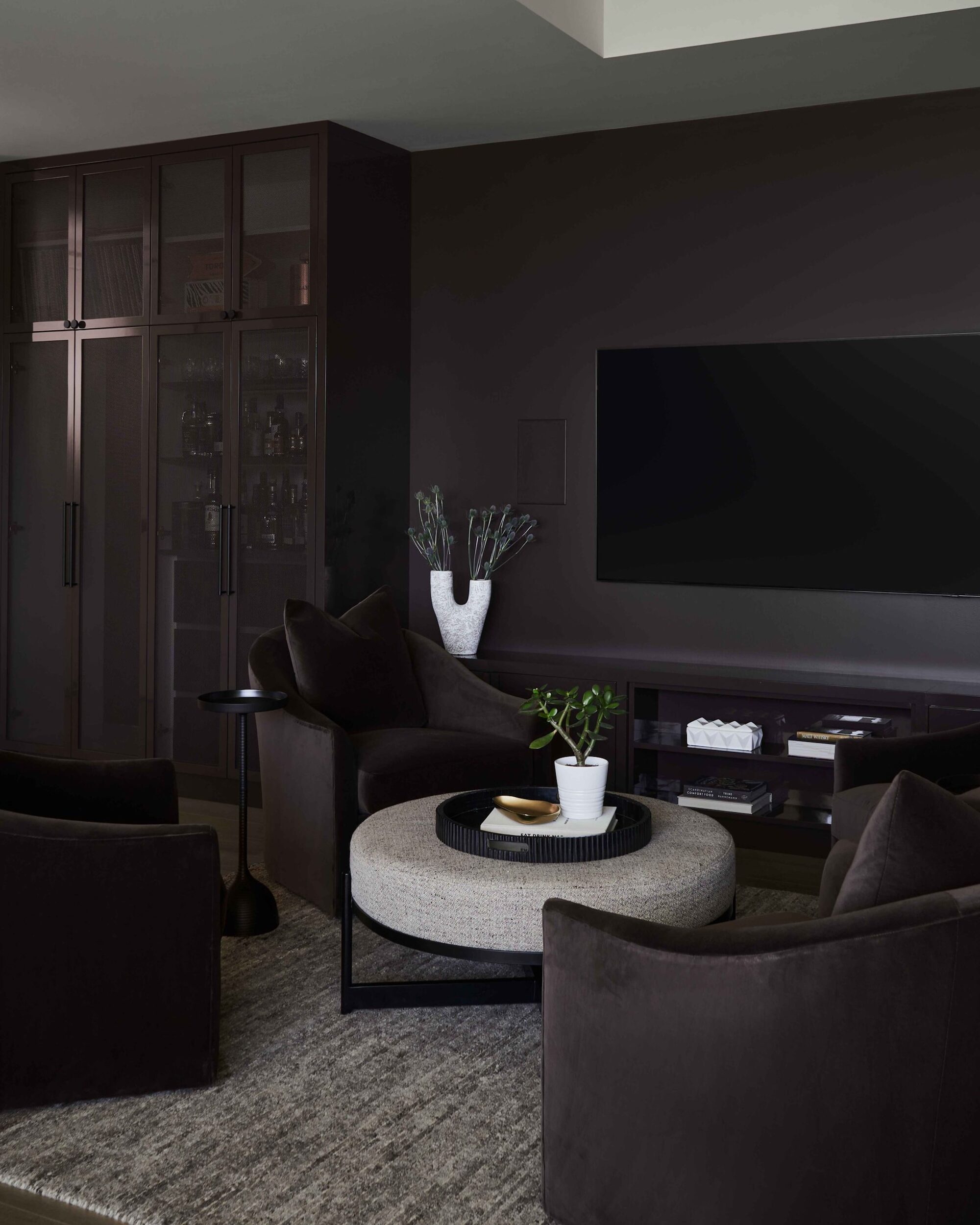 dark living room with black walls, black mesh cabinetry, black velvet armchairs around a gray upholstered round coffee table, on a gray marle rug