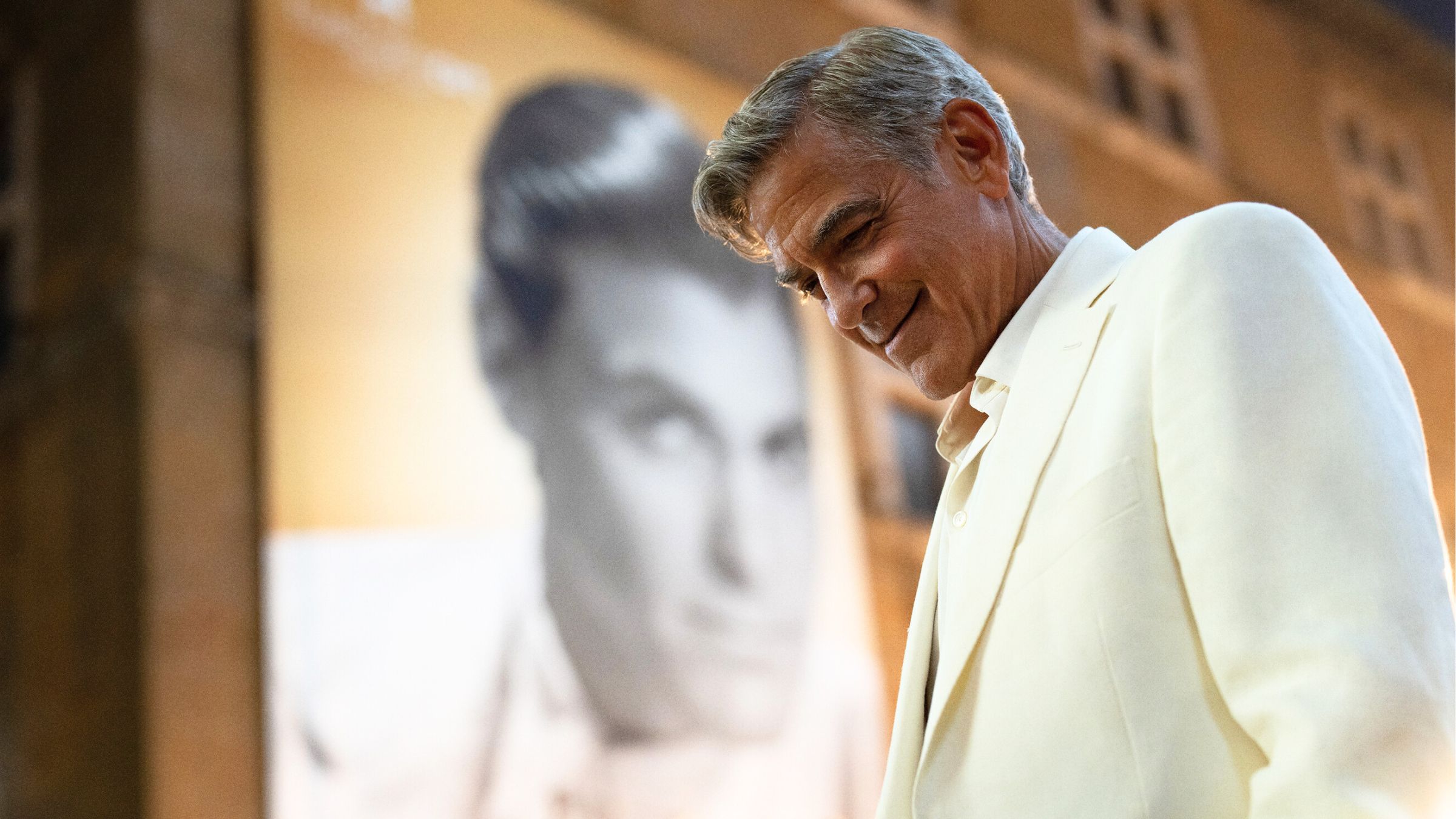 George Clooney smiles with his head hung low in front of a black and white poster of himself