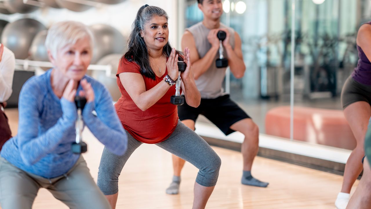 In a group fitness class, three people hold a dumbbell to their chest and perform a goblet squat, with their feet and knees wide. 