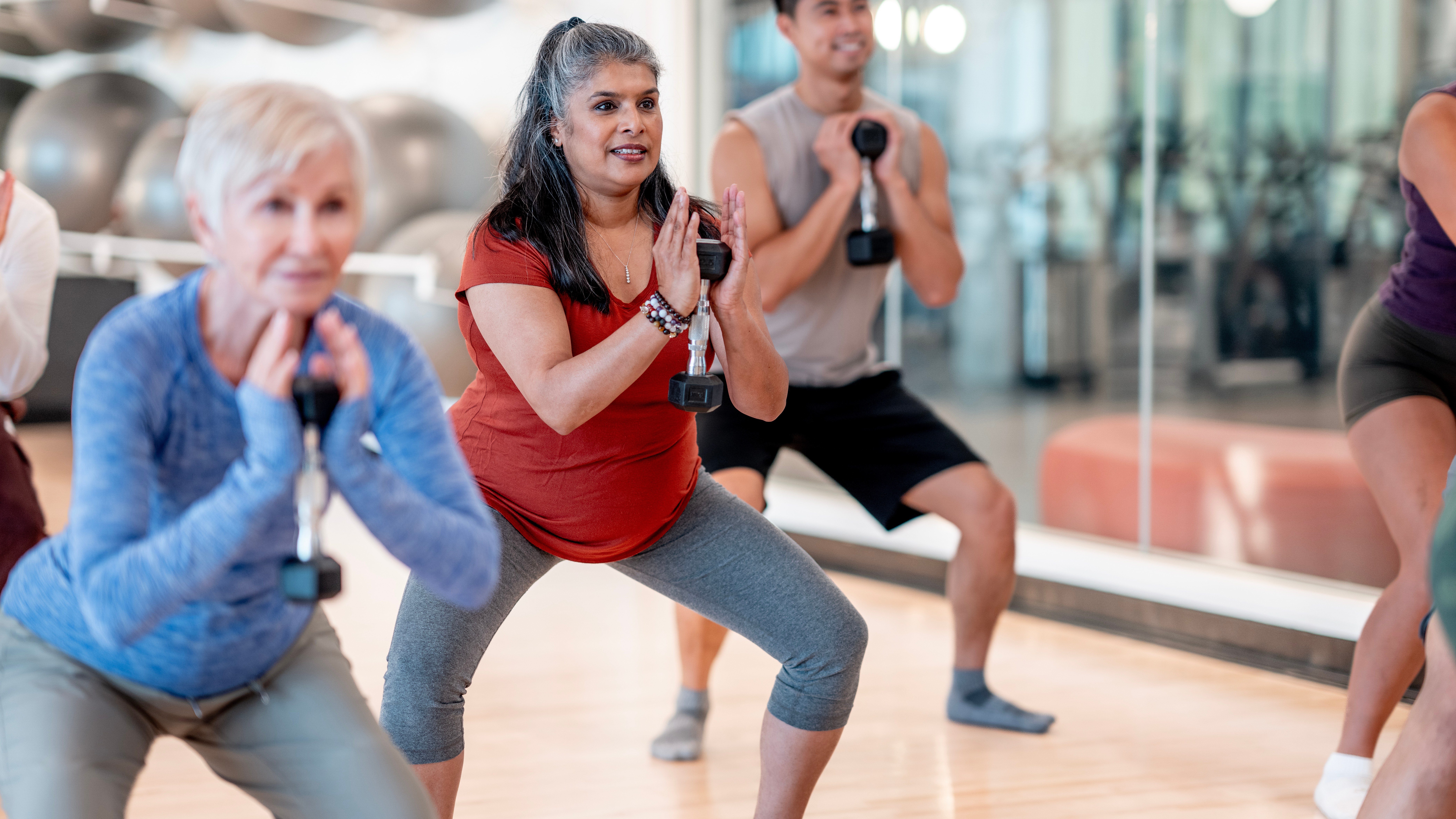 In a group fitness class, three people hold a dumbbell to their chest and perform a goblet squat, with their feet and knees wide. 