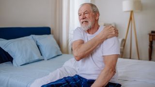 Man holds his shoulder while sitting on the edge of his bed