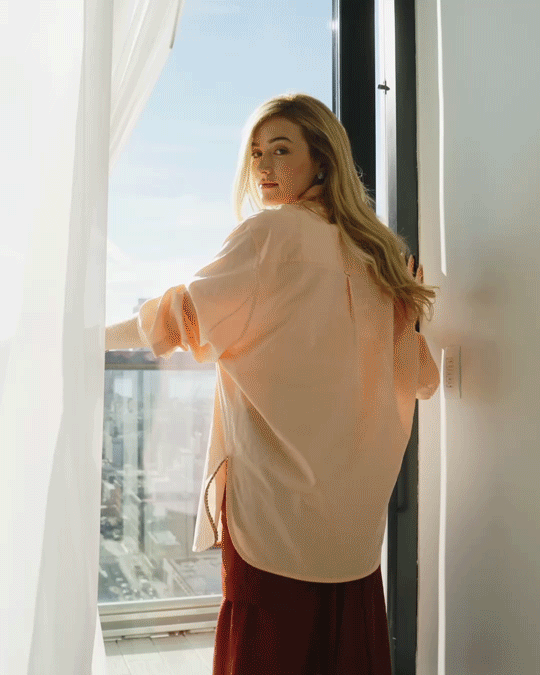 gif of Sarah Pidgeon standing by a window wearing an apricot shirt and red skirt