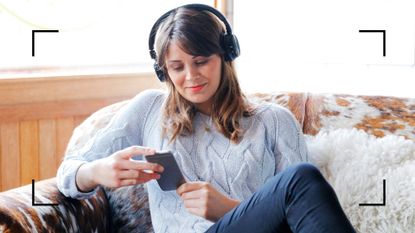 Woman wearing over-ear headphones listening to the best relationship podcasts, holding phone and sitting on armchair