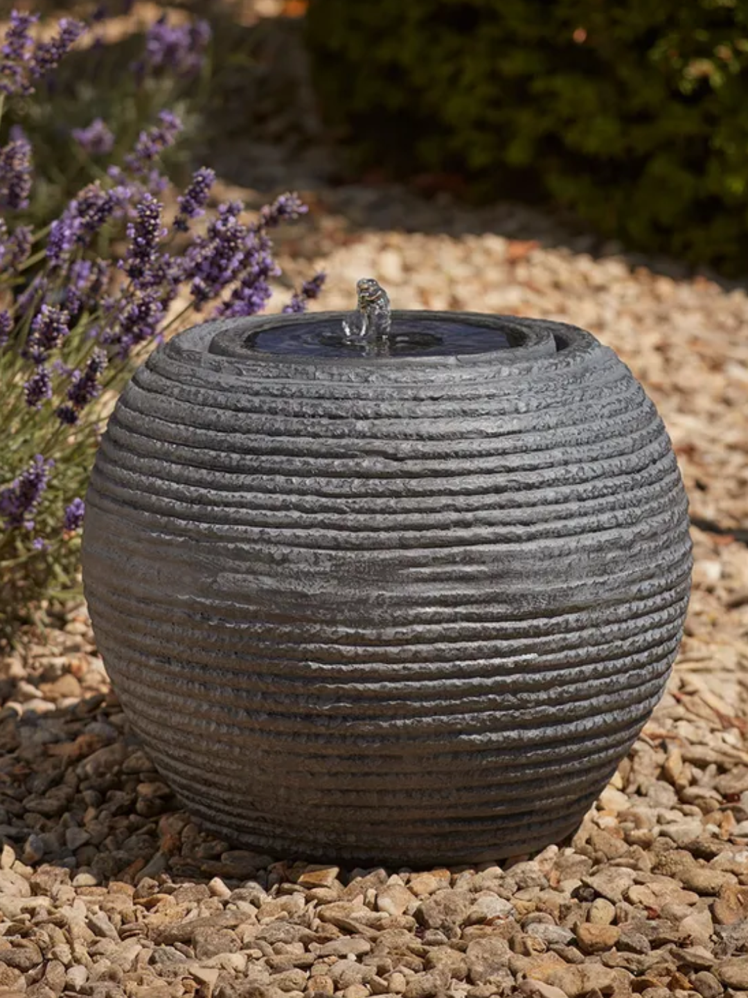 An outdoor dark gray fountain on a gravel path by lavender
