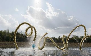 Daytime, outside image, close up a gold and glass decorative piping garden water feature, trees in the backdrop, blue cloudy sky