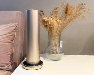 A sleek, cylindrical, brushed metallic gold essential oil diffuser on a white table next to a clear ribbed vase of dried florals and a pale pink sofa