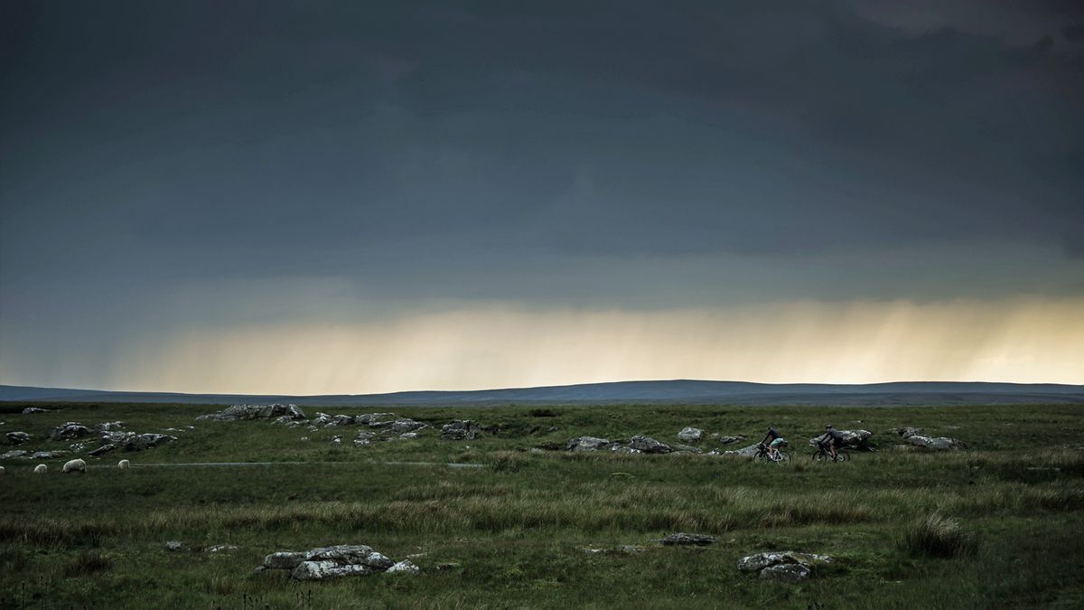 Two cyclists ride with storm clouds in the distance