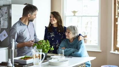 Woman with arm around mature woman looking towards man in kitchen