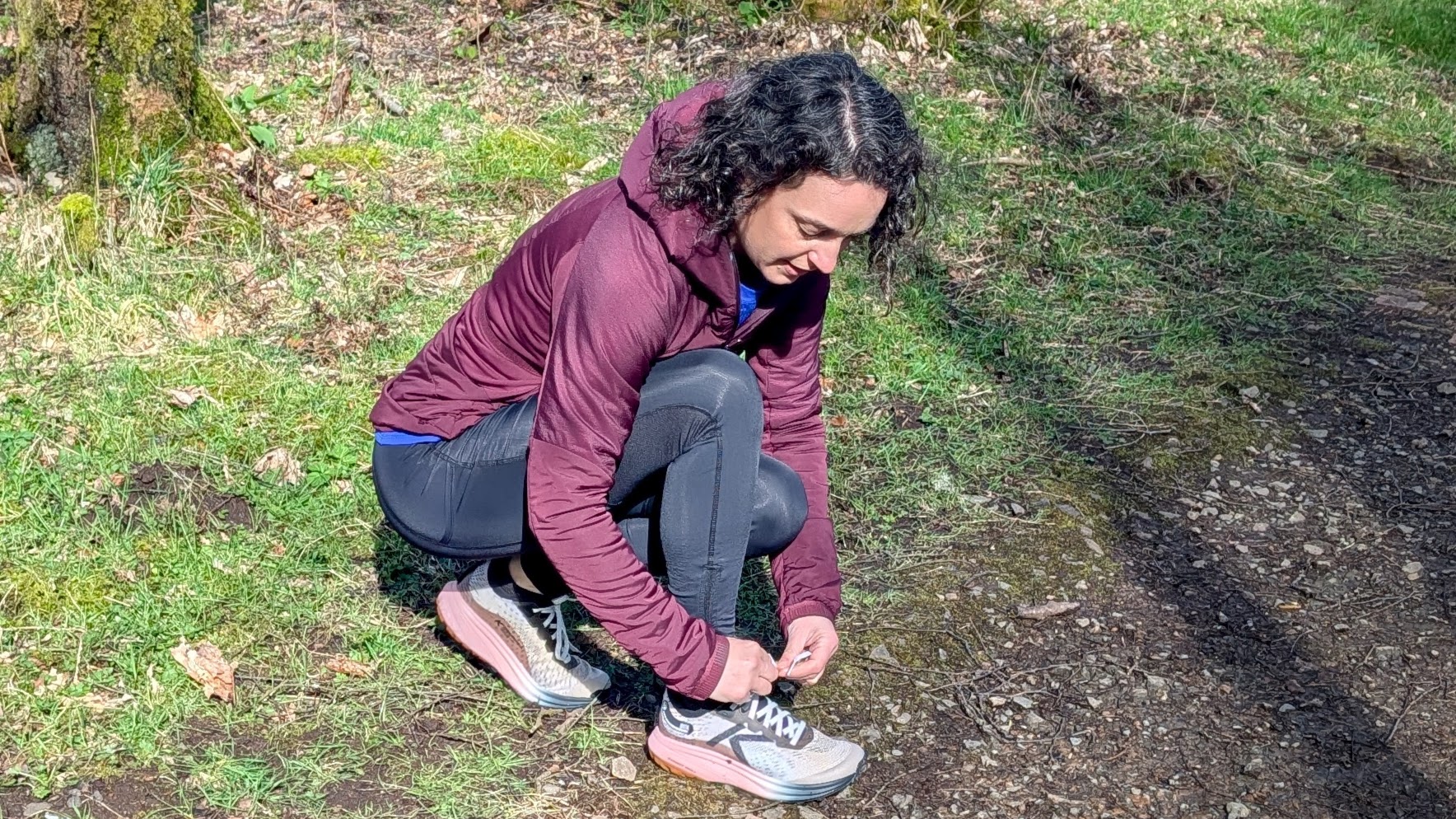 A runner in a maroon jacket tying the laces on her Keen Seek trail running shoes