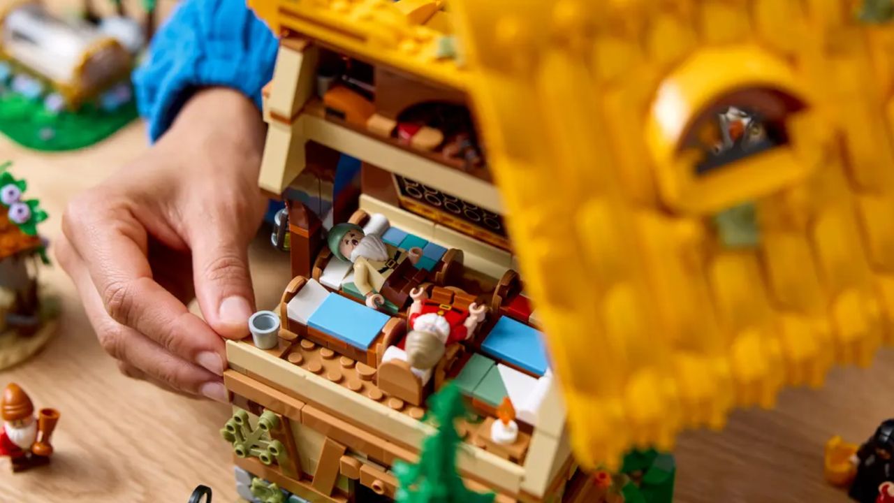 A hand moves the Lego Snow White and the Seven Dwarfs' Cottage set on a wooden table, with minifigures inside