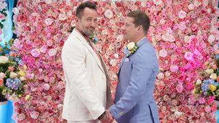 John Paul and Jez wearing wedding suits and holding hands in front of a wall of roses.