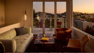 A living room at sunset with views over San Fransisco bay from bifolding doors and a full wall window with a green sofa and two orange mis-matched accent chairs