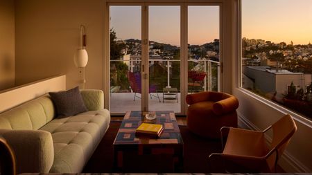 A living room at sunset with views over San Fransisco bay from bifolding doors and a full wall window with a green sofa and two orange mis-matched accent chairs