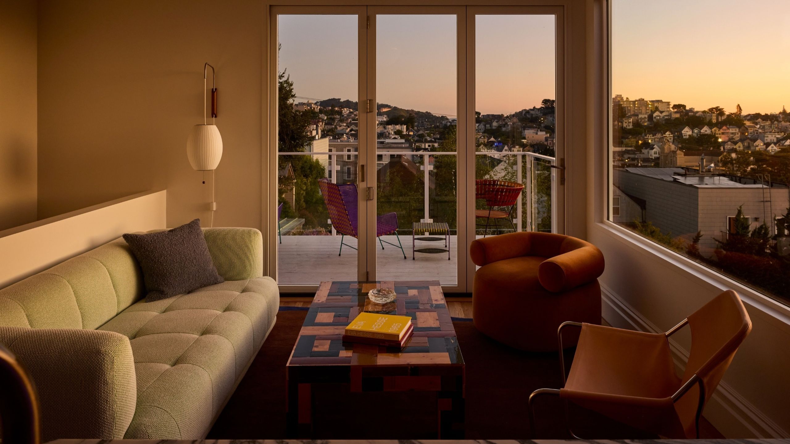 A living room at sunset with views over San Fransisco bay from bifolding doors and a full wall window with a green sofa and two orange mis-matched accent chairs