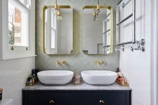 Dark blue vanity unit with twin sinks, brass fittings, brass mirrors and pastel green metro tiles in a herringbone pattern