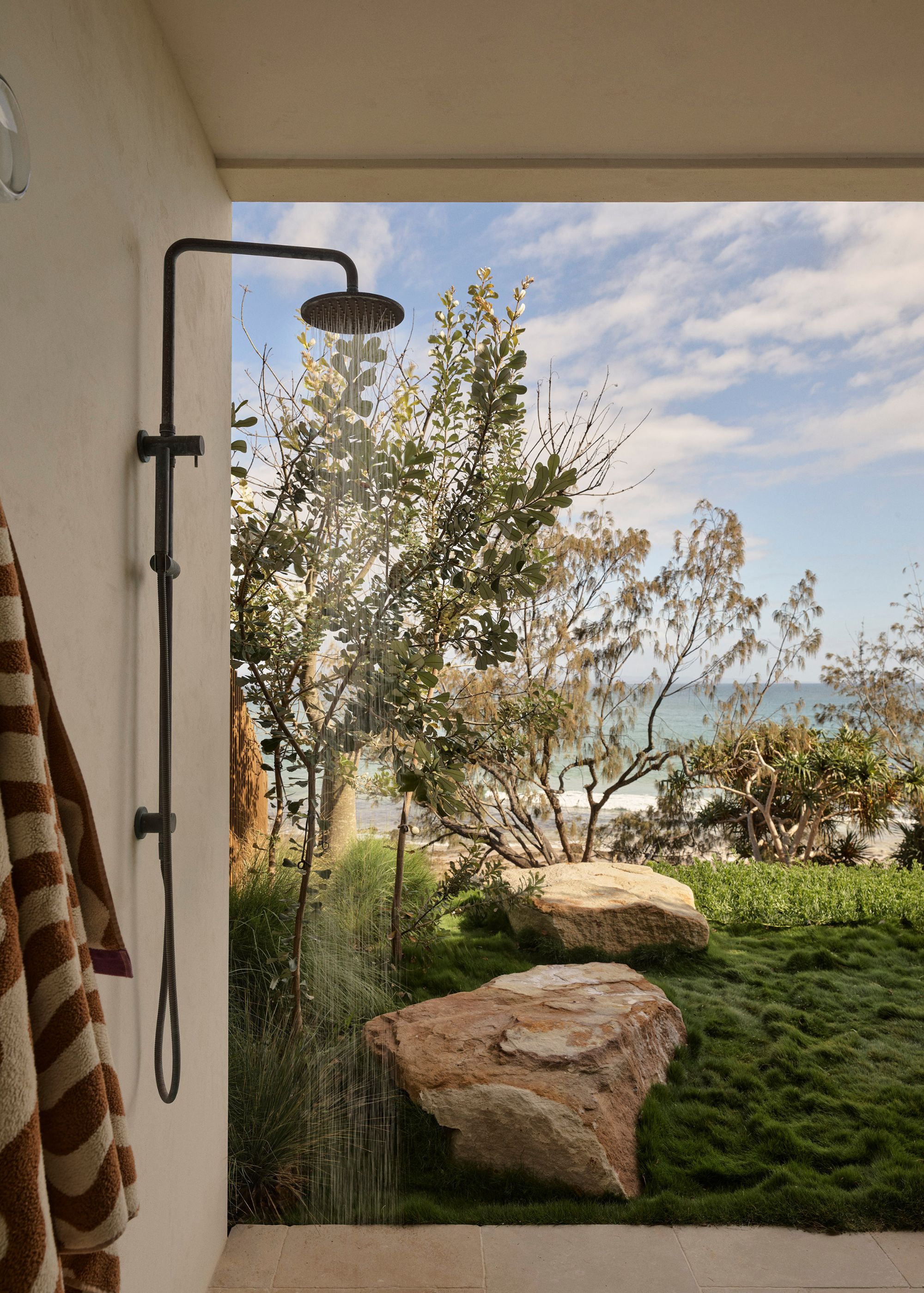 An outdoor shower facing a rock garden with a lush lawn and tall trees