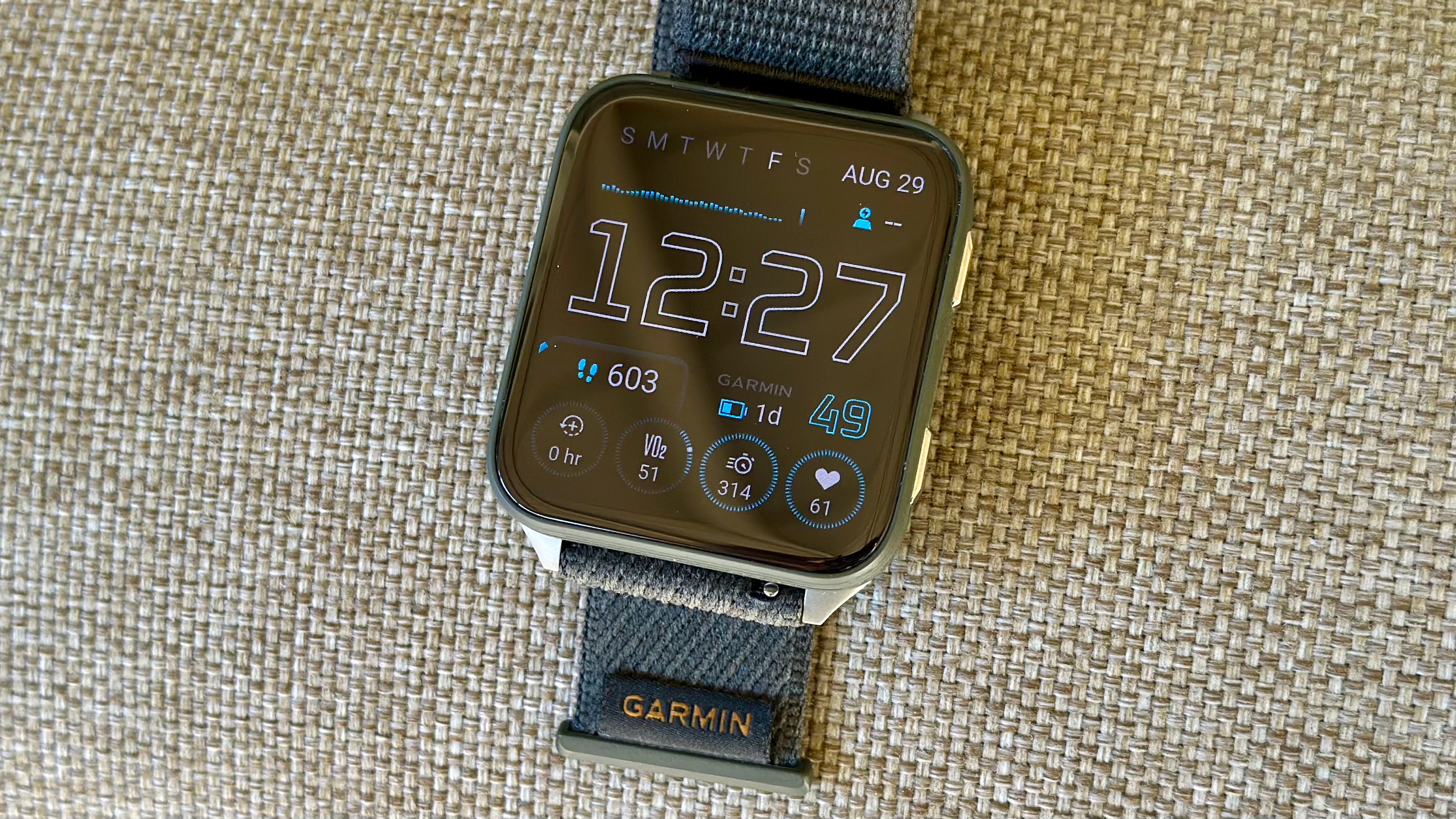 The Garmin Venu X1 sitting on a chair back, showing the AOD mode for the default Snapshot watch face showing a variety of data fields, including HR, stress, steps, battery, VO2 Max, intensity minutes, and recovery time, along with the time and date.
