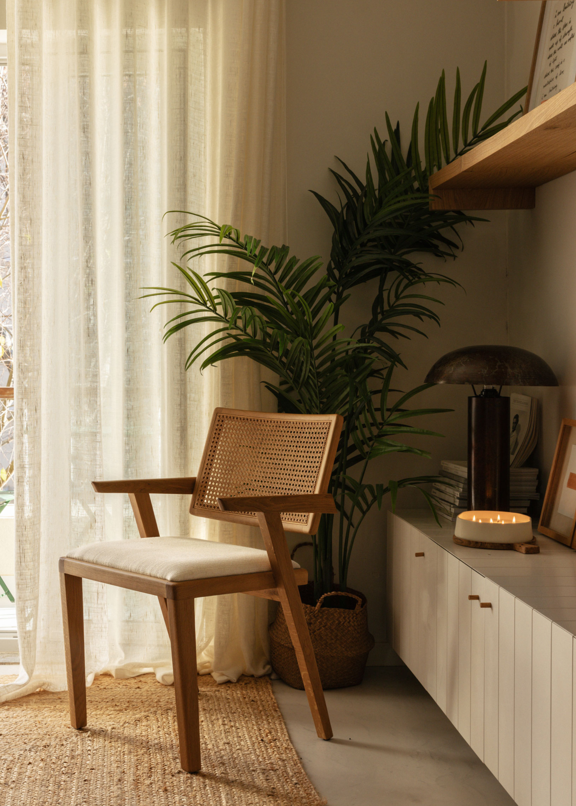A living room corner with a rattan chair, a potted plant, a white sideboard with a black lamp, a multi-wick candle and leaning art