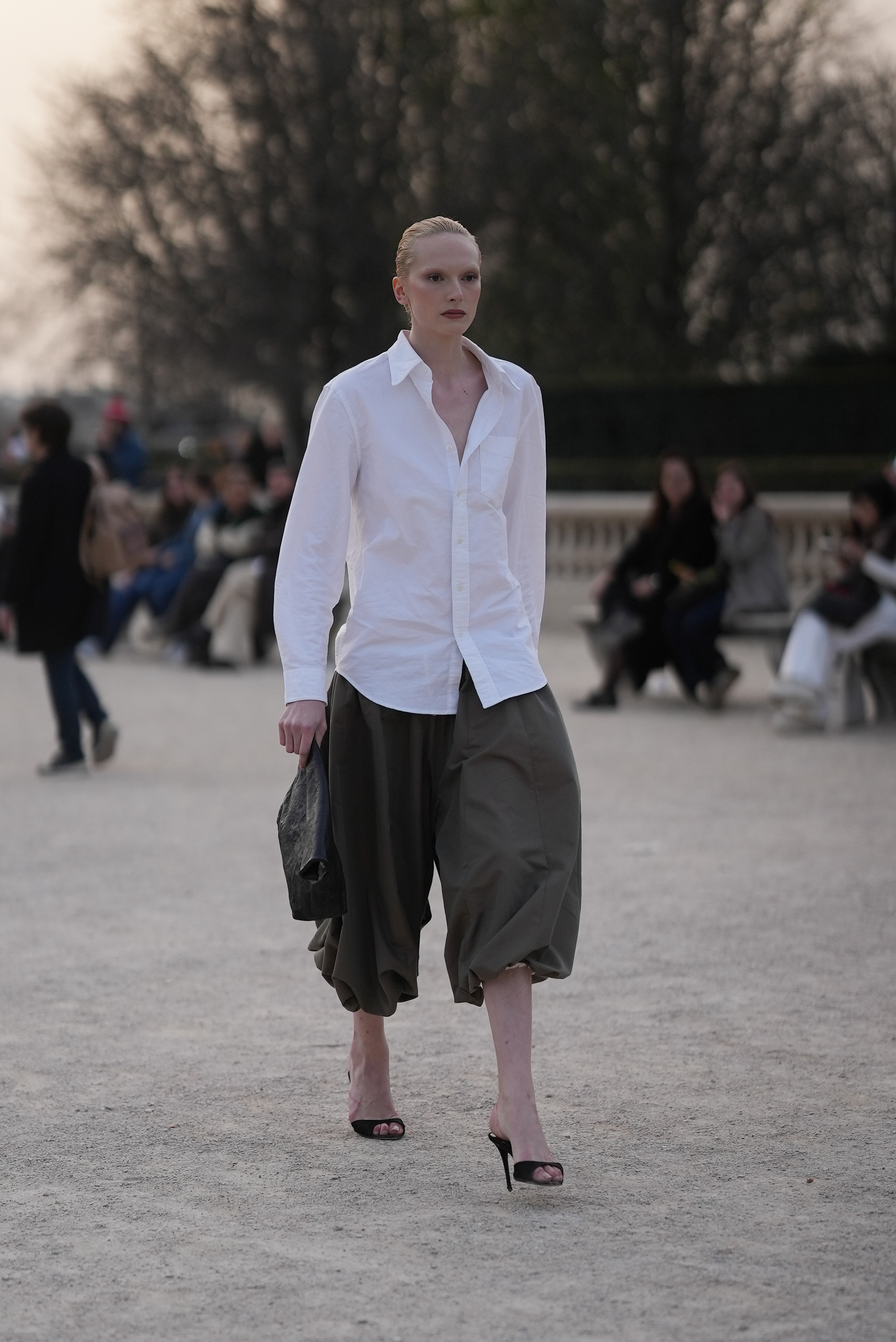 Alex Consani is wearing a white oversized cotton button down shirt, khaki-green wide leg capri harem trousers in a balloon-shaped leg; black open toe mule heels with a peep toe a leather clutch.