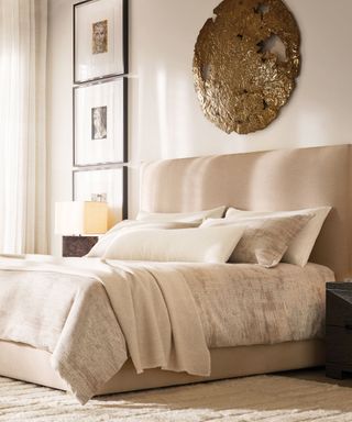 White and beige bedding on a beige bed frame against a white wall, decorated with three portrait photographs and gold art.