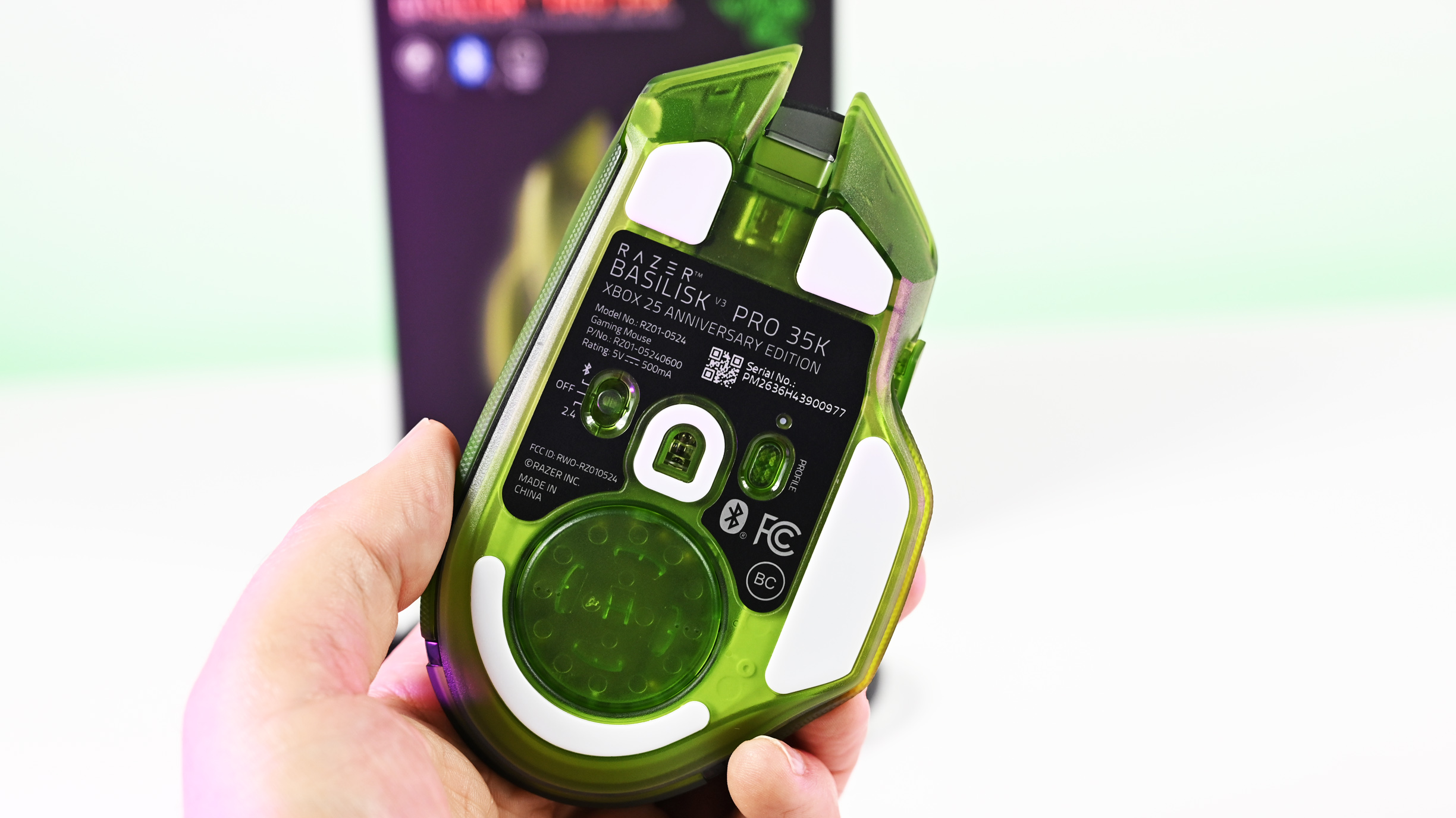 Underside of the Razer Basilisk V3 Pro 35K Xbox 25th Anniversary mouse held in hand, showing white PTFE feet, optical sensor, and power switch.