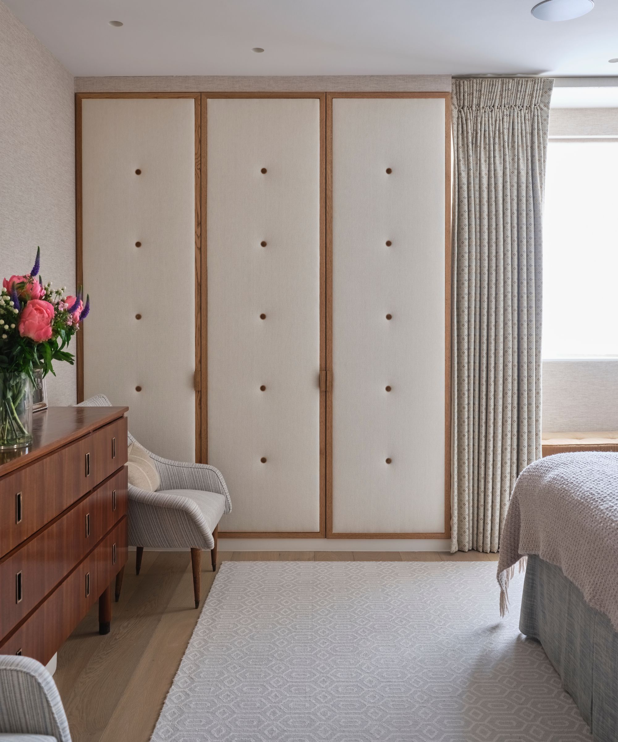 A cream and wood padded closet in a bedroom with a cream rug, wooden sideboard with vase of bright pink flowers, and grey chair.
