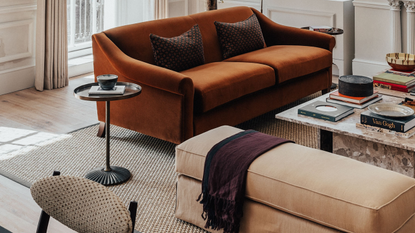 modern living room with orange sofa, long ottoman seat, side table, square marble coffee table styled with books, a potted plant in the corner, sash windows with curtains, decorative cornice, and a jute rug