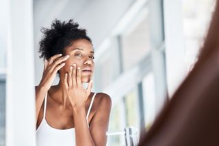 A woman looking in the mirror while touching her face inquisitively, to illustrate what are acne scars, and what you should do to prevent them
