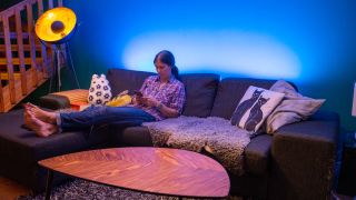 Nanoleaf Essentials Lightstrip lyser i blått bakom en soffa där en kvinna sitter och flippar på mobilen.