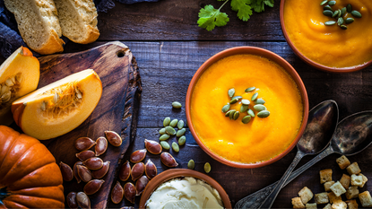 Two bowls of pumpkin soup sit on wooden table, surrounded by ingredients 