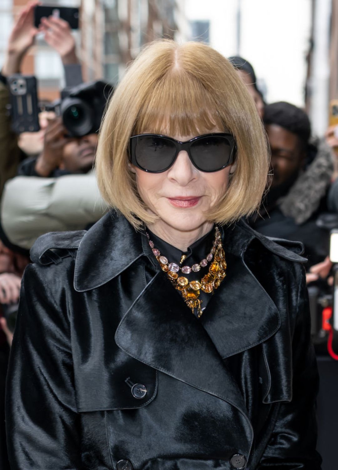 Anna Wintour in a black coat and oversized sunglasses with amber jewelled necklace
