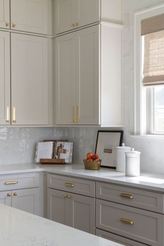 Image of a white kitchen. The backsplash is made of white zellige tile, and the cabinets are a light gray-beige. On the cabinetry there are gold cabinet pulls with matching gold cabinet backplates.