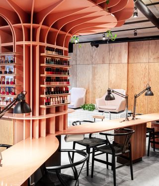 Interior view of Le Hideout nail bar featuring a large peach coloured structure with shelves and nail polish, wood covered walls, grey floors, pink and black chairs, manicure stations, black lamps and green plants