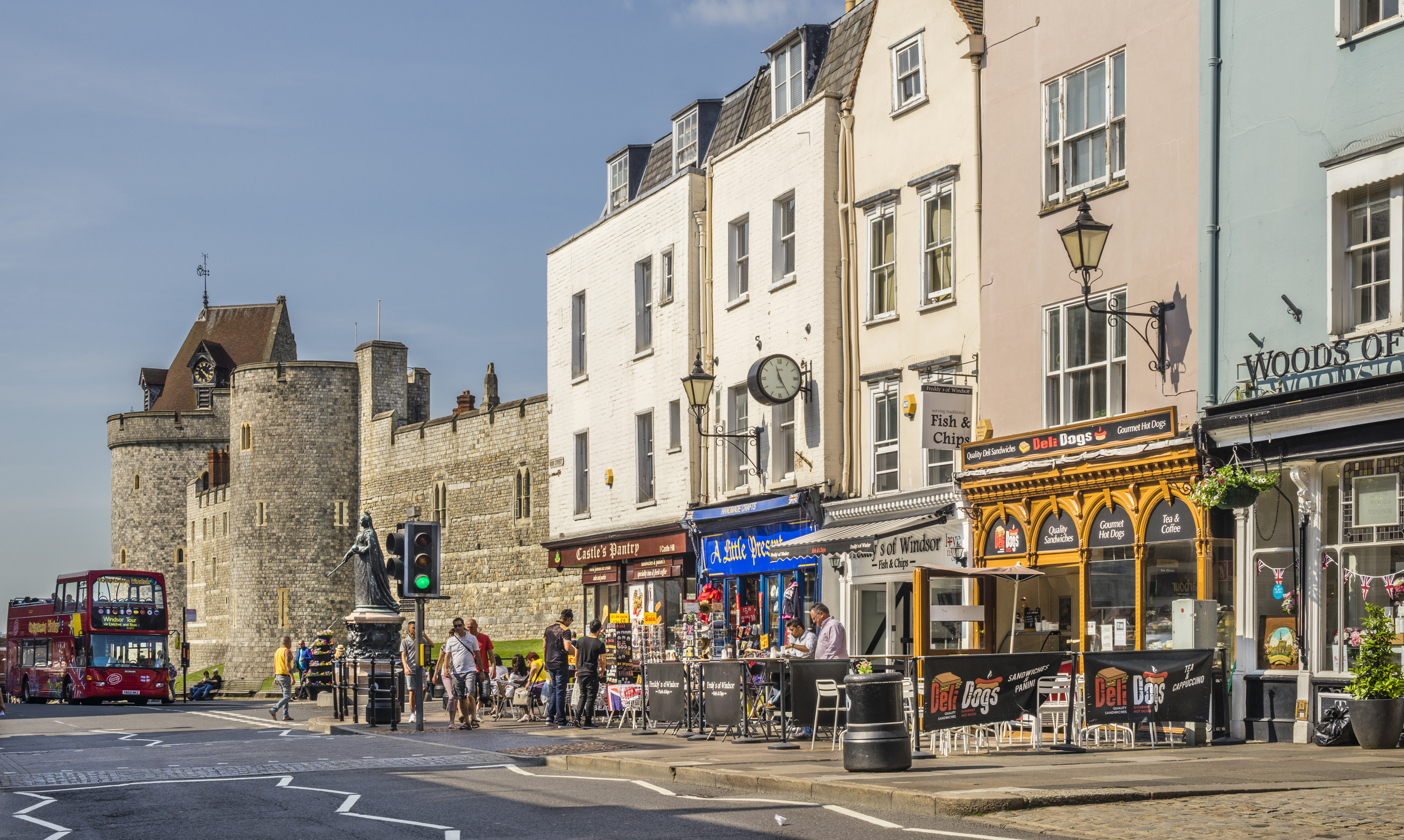 Small business shop fronts on Windsor High Street with Queen Victoria statue and the Lower Ward of Windsor Castle with shoppers and pedestrians