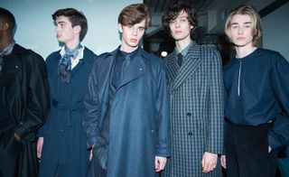 Four male models wearing suits and trousers in shades of blues and blacks, raglan-sleeved trenches, complete with exaggerated epaulets, which were teamed back with Shetland wool crew neck sweaters and loosely cut trousers, finished with pussy-bow tied fringed scarves.