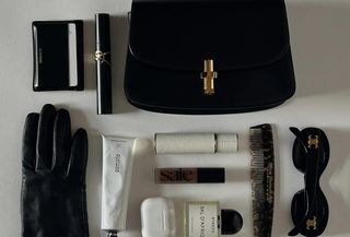 a close-up photo of a woman's bag contents laid out with one of the best wallets for women, along with mascara, gloves, perfume, foundation, a comb, sunglasses and headphones