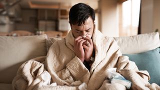 A young man is shown sat on a couch with a blanket wrapped around him. He is blowing into a tissue. 