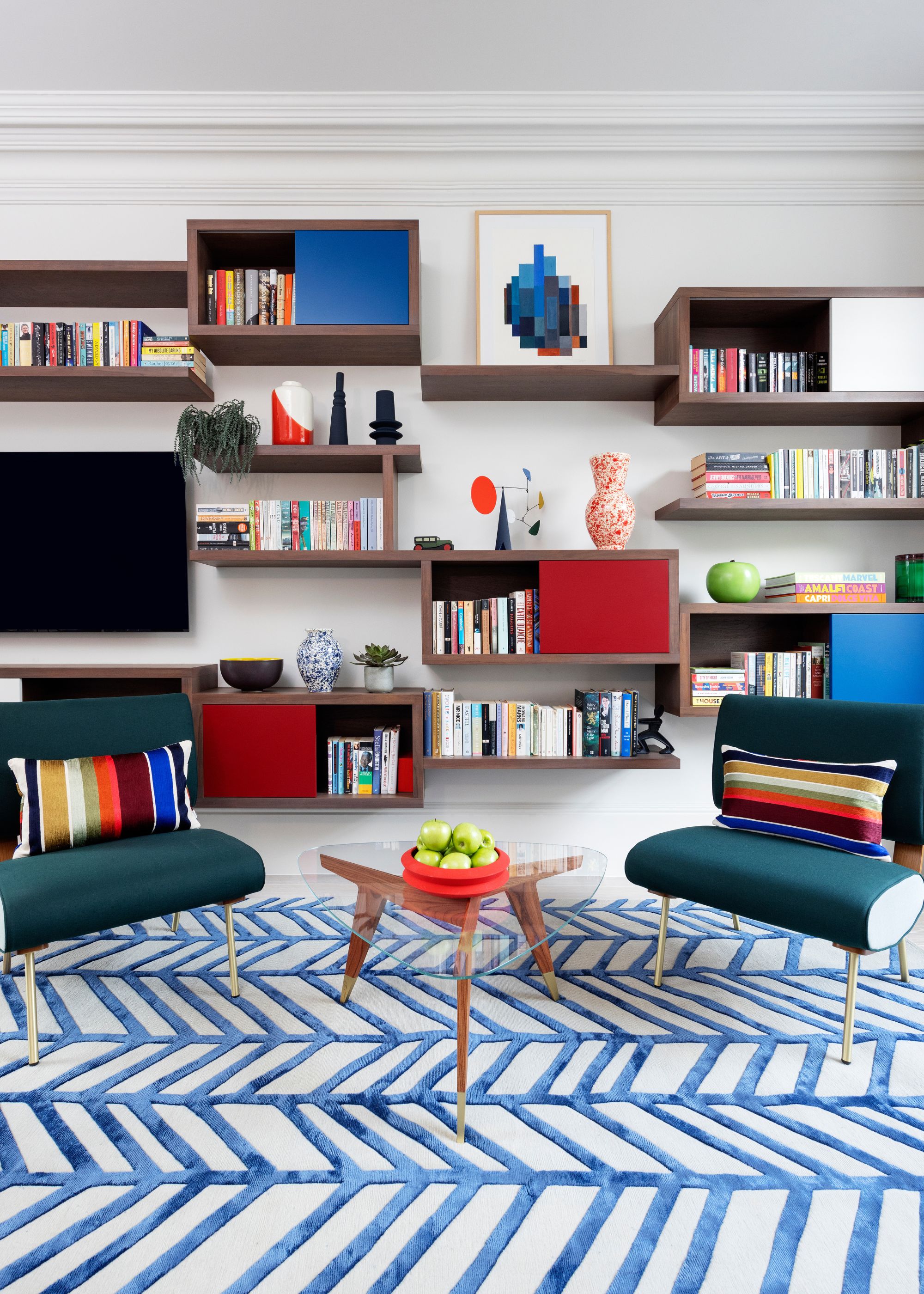 A colorful living room with a bespoke media wall featuring a TV and open shelving. Two teal accent chairs and a blue and white geometric rug on the ground.
