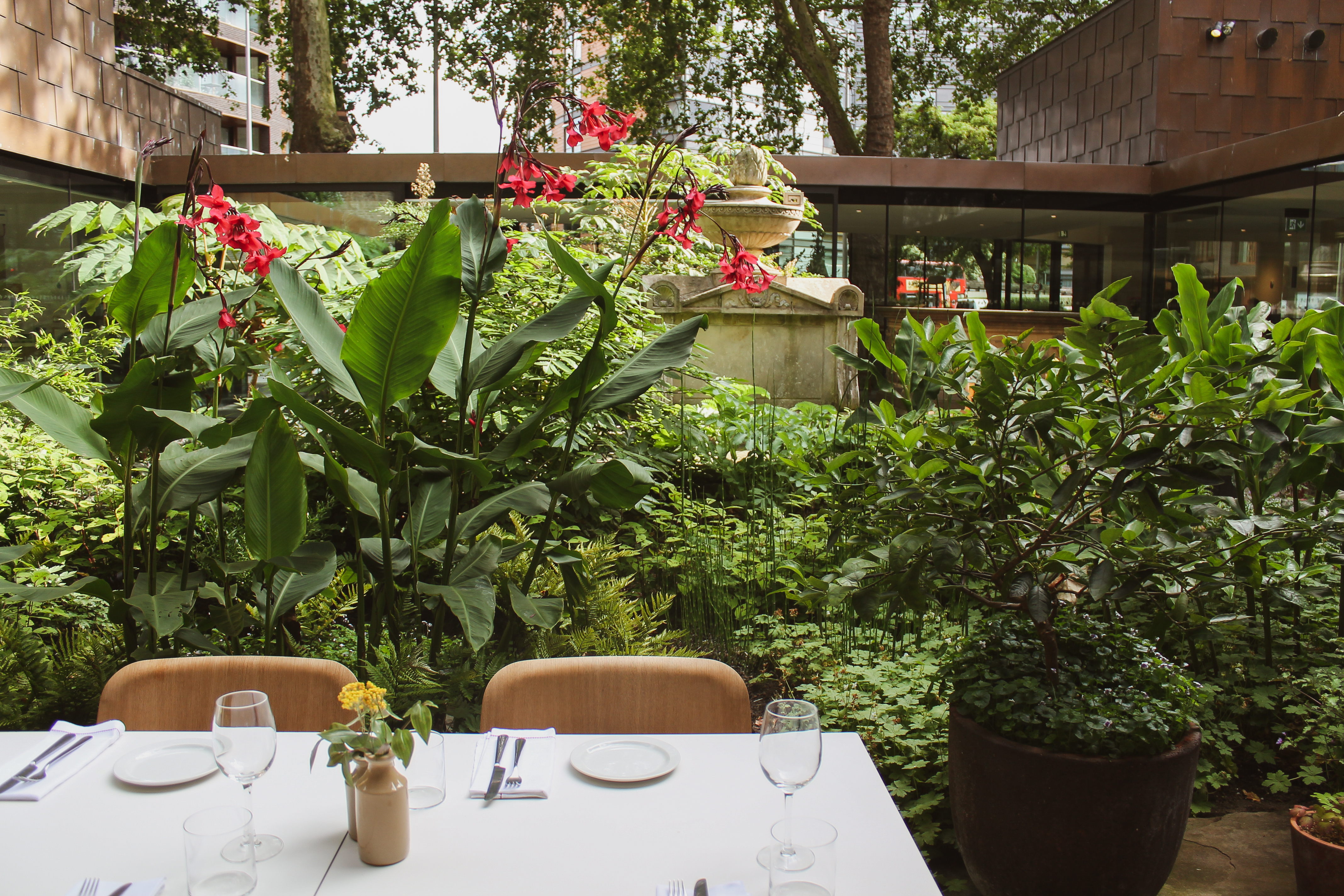 A table with white tablecloth and two pale wooden chairs, outside, with a very leafy background.
