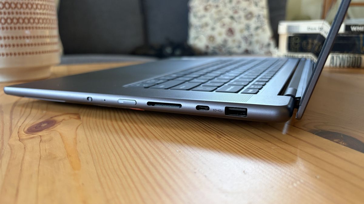 A slim silver laptop rests open on a wooden table, with its keyboard and side ports in focus; the cozy home setting feels calm and practical.