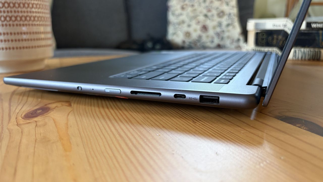 A slim silver laptop rests open on a wooden table, with its keyboard and side ports in focus; the cozy home setting feels calm and practical.